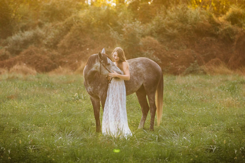 girl in white dress holding face of grey pony in field