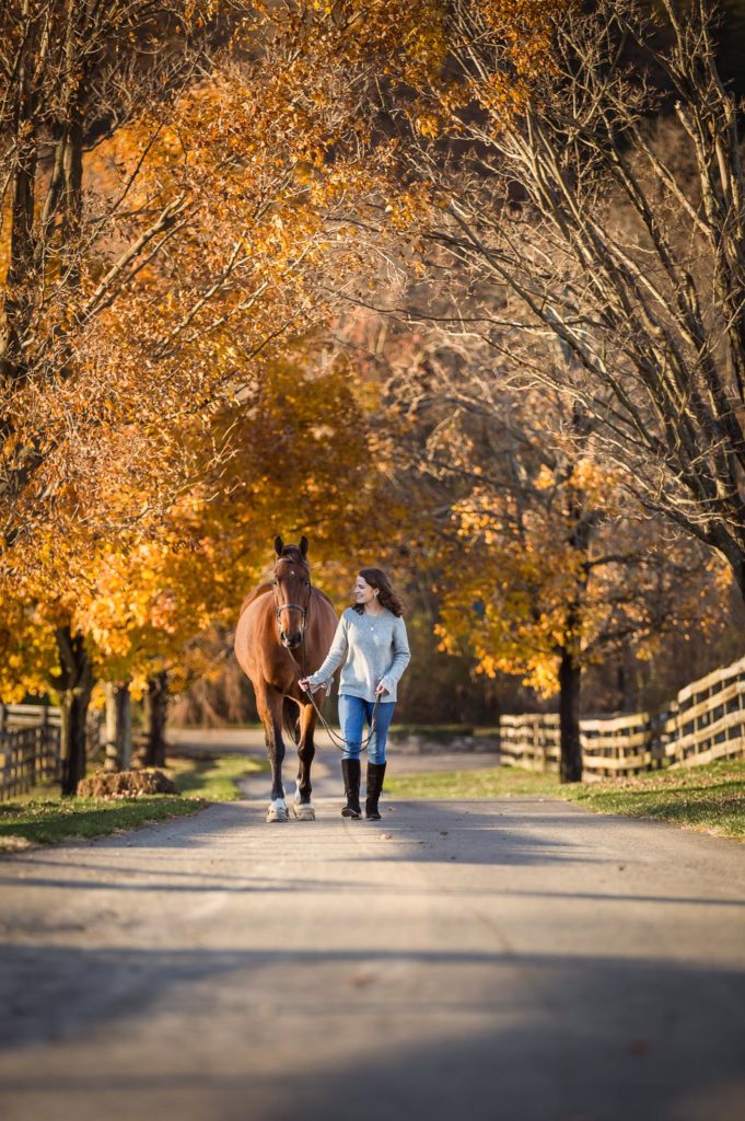 girl walking horse through fall trees at Hunters Crossing Farm in Long Valley, NJ