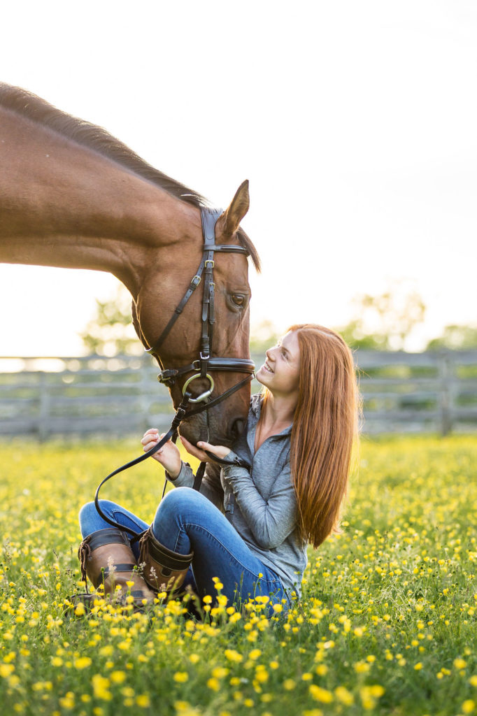 red headed girl sitting in field of buttercups with her horse