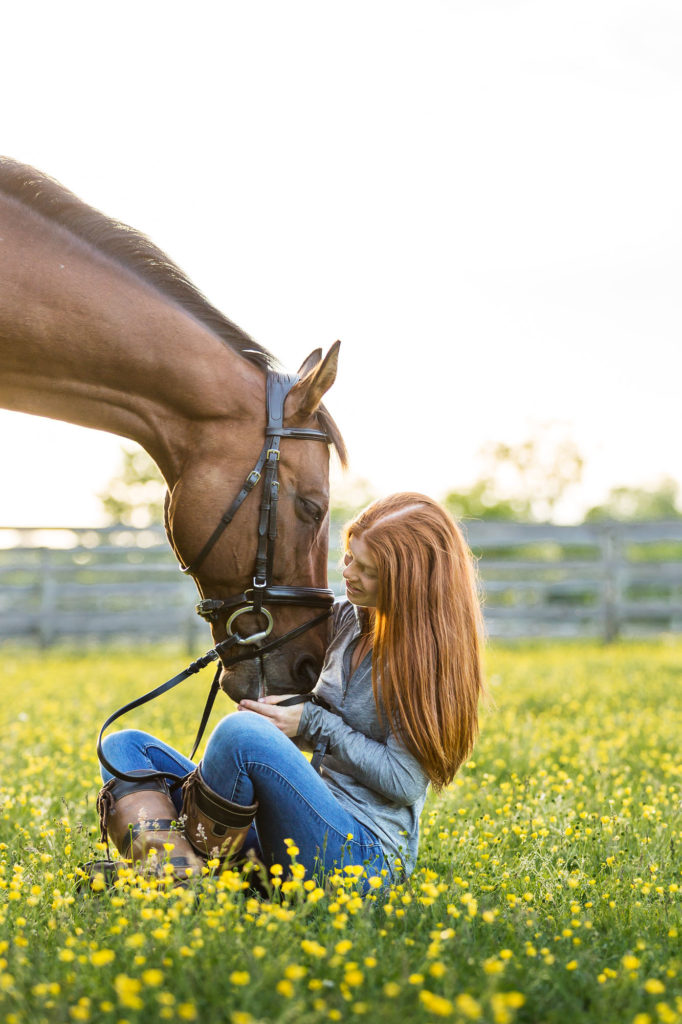red headed girl sitting in field of buttercups with her bay OTTB at Golden Gait Farm