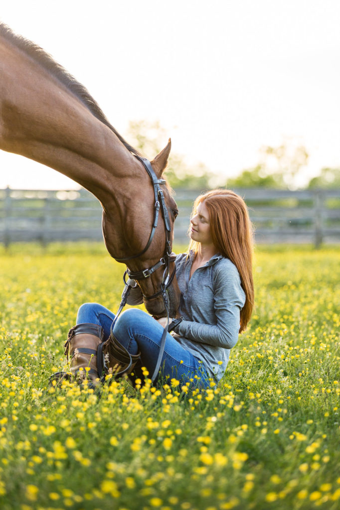 red headed girl sitting in field of buttercups petting her horse