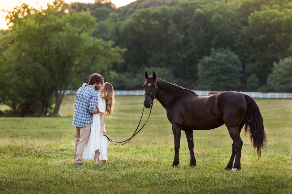 young couple kissing with black horse in field at Great Scott Farm