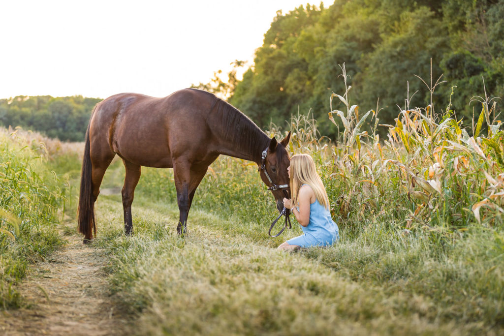 woman in blue dress sitting on ground kissing her horse in corn field